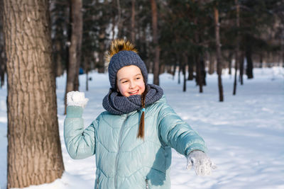 A cheerful girl in warm clothes throws a snowball in a winter park. outdoor games.