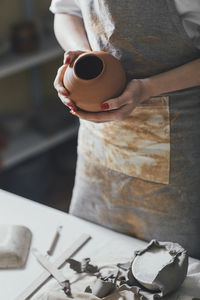 Midsection of female potter holding pot while standing by table in workshop