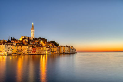 Illuminated buildings by sea against sky in city