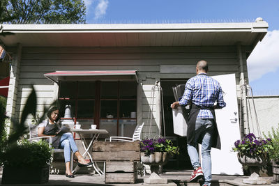 Man carrying serving trays while walking by woman working at table outside cafe