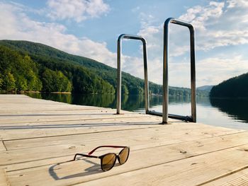 Black sunglasses on wooden pontoon near a lake surrounded by green forest