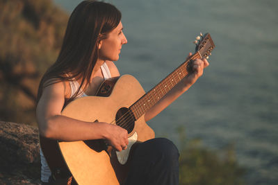 A beautiful woman playing acoustic guitar sitting on the rock. pretty girl practicing music guitar