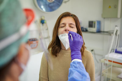 Back view of anonymous medic in protective shield taking analysis from nose of female patient while using medical tool during covid 19 pandemic