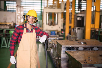 Smiling asian factory young worker with plaid red shirt, helmet, safety glasses at trolley