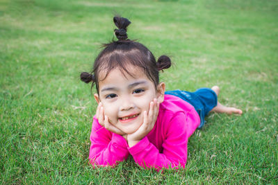 Portrait of smiling cute girl with hand on chin lying on grass at park