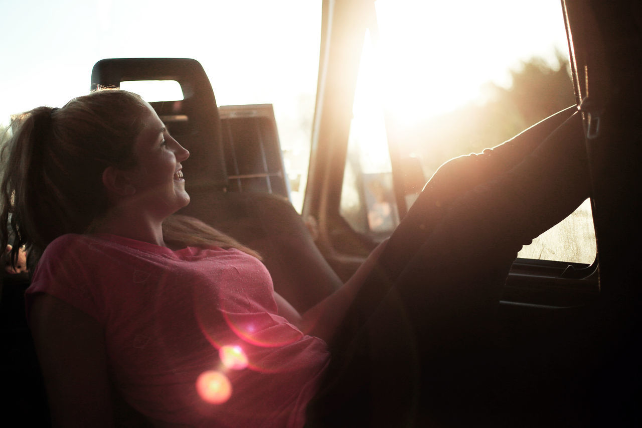 mode of transportation, vehicle interior, women, real people, leisure activity, lifestyles, travel, sitting, transportation, journey, land vehicle, motor vehicle, car, window, adult, one person, relaxation, day, sunlight, outdoors, lens flare, hairstyle, road trip