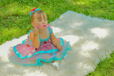 Full length of cute girl blowing bubble wand while sitting on rug at field