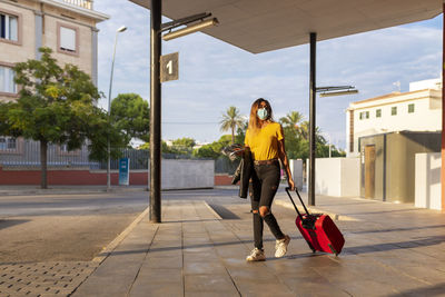 Young woman wearing protective mask while walking with luggage at bus stop