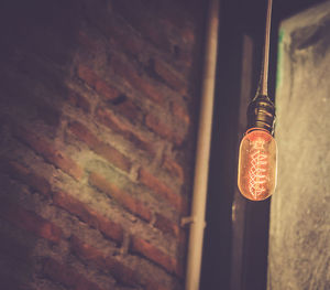 Low angle view of illuminated light bulb hanging by brick wall