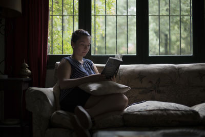 Woman reading book while sitting on sofa at home