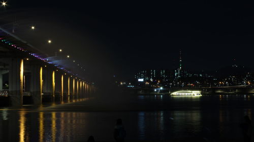 Illuminated bridge over river by buildings against sky at night