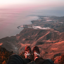 Low section of man sitting on cliff against sea during sunset