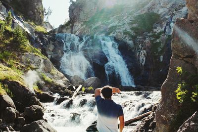 Rear view of man standing against waterfall during sunny day