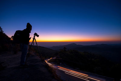 Side view of man with camera and tripod standing on mountain against sky during sunset