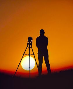 Silhouette man standing by camera on land against orange sky during sunset