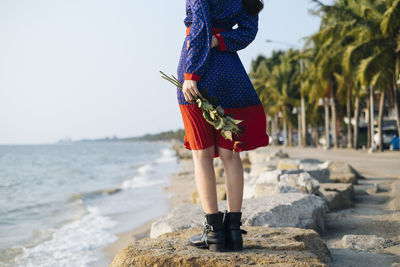 Low section of woman standing on rock by sea shore with rose in hand