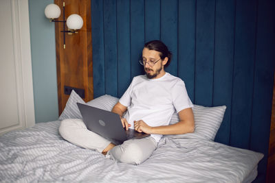 Man in a white t-shirt, a freelancer with glasses, sits on the bed at home with a laptop and works 