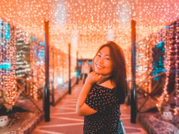 Portrait of smiling young woman standing against illuminated lights