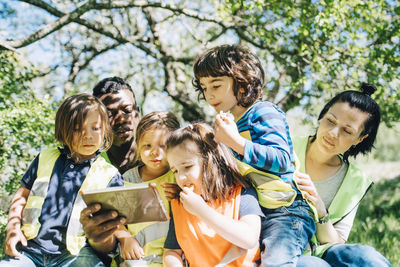 Multi-ethnic students and teachers sharing digital tablet while sitting against trees in playground