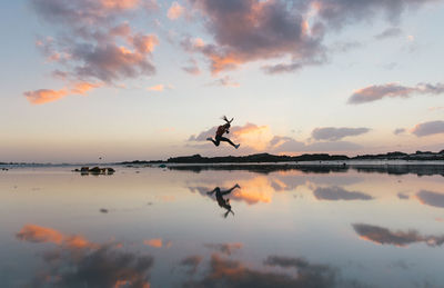 Woman jumping over lakeshore against sky during sunset