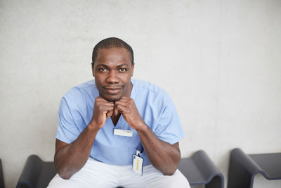 Portrait of confident mid adult male nurse sitting against wall at hospital