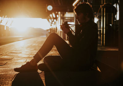 Side view of boy holding camera sitting at railroad station platform