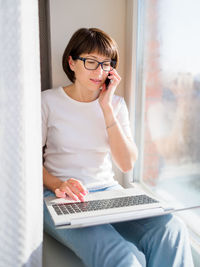 Woman with smartphone remote works from home. she smiles widely on window sill with laptop on knees.