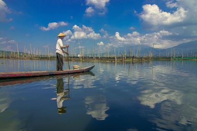 Man standing on rowboat in lake against sky