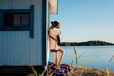 Woman stood leaning against a wooden hut whilst playing the guitar