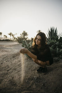 Young woman playing with sand while crouching by cactus plant