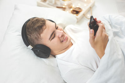  teenage boy in a white t-shirt lies in bed with headphones and a smartphone in his hands. 