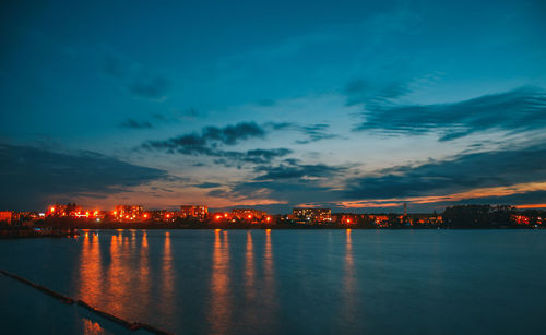 Illuminated city by sea against sky at sunset