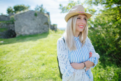 Smiling woman in sun hat with arms crossed standing on grassy field during sunny day