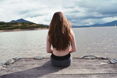 Rear view of woman sitting by lake against sky