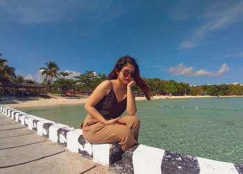 Portrait of young woman sitting on wall by sea