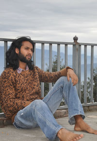 A long haired and bearded young guy looking sideways while sitting with leaning on safety barrier