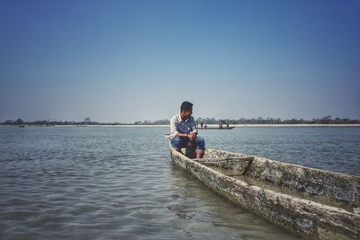 Man sitting on riverbank against clear blue sky