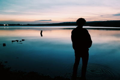 Silhouette boy standing by lake against sky during sunset