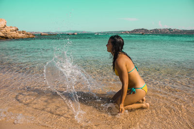 Side view of young woman in bikini kneeling at beach