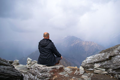Rear view of mid adult man sitting on mountain against cloudy sky