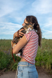 Woman pet owner holding and hugging domestic cat in hands walking outside among countryside