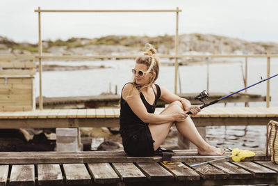 Mature woman with fishing rod looking over shoulder while sitting on jetty at harbor