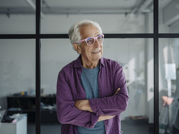 Smiling entrepreneur wearing pink eyeglasses looking away while standing with arms crossed at office