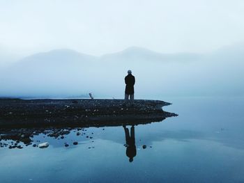 Man standing by lake overlooking misty mountains