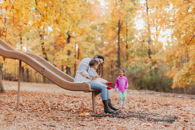Full length of mother sitting on slide with kids at park during autumn