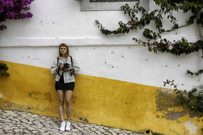 Full length of woman holding camera standing against wall