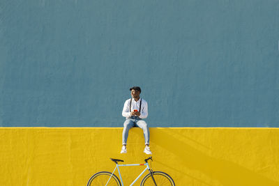 Young man with mobile phone and bicycle sitting on yellow wall