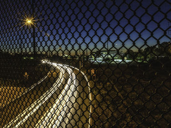 Chainlink fence by illuminated street against sky at night
