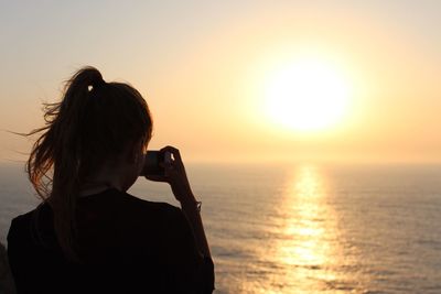 Woman photographing sea against sky during sunset