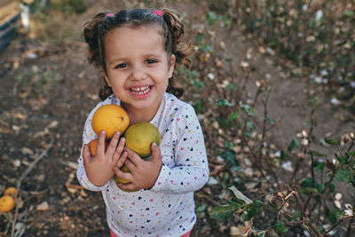 From above of carefree little kid standing with pile of fresh oranges in countryside and looking at camera